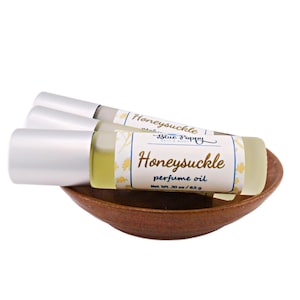 May include: Three silver and glass roller bottles with a yellow label that reads "Honeysuckle perfume oil Net Wt. .30 oz / 8.5 g" and a blue label that reads "Made by Blue Poppy Oils". The bottles are sitting in a brown wooden bowl.