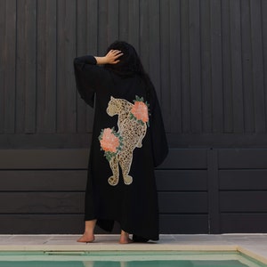 May include: A black kimono robe featuring a large graphic print on the back. The design depicts a spotted leopard in tan and brown tones, surrounded by large, vibrant coral-pink flowers with green leaves. The robe has wide, loose-fitting sleeves.