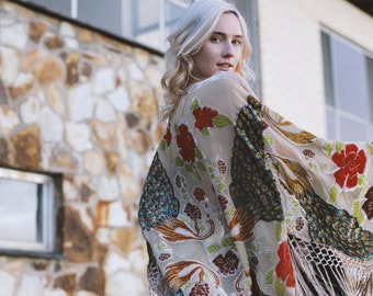 May include: A woman with blonde hair wearing a long, flowing kimono with a floral and peacock design. The kimono is beige with red, green, and blue accents. The kimono has fringe along the bottom.