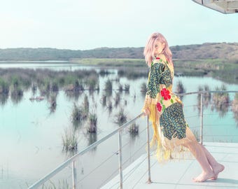 May include: A patterned kimono with red floral details and fringing. The garment is worn outdoors near a lake. The background shows a tranquil landscape with water and marsh vegetation.