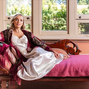 Op de afbeelding: Een vrouw in een witte satijnen slipjurk en een bordeauxrode fluwelen sjaal met bloemenborduursel zit op een chaise longue. Ze houdt een roze bloem in haar hand. De chaise longue heeft een roze fluwelen kussen en sierlijke houten houtsnijwerk.