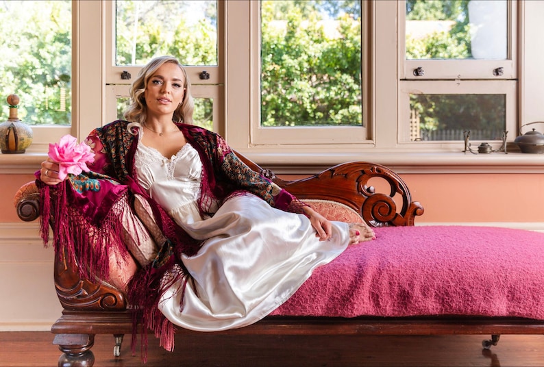 Op de afbeelding: Een vrouw in een witte satijnen slipjurk en een bordeauxrode fluwelen sjaal met bloemenborduursel zit op een chaise longue. Ze houdt een roze bloem in haar hand. De chaise longue heeft een roze fluwelen kussen en sierlijke houten houtsnijwerk.