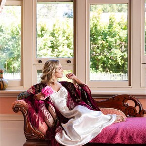 Op de afbeelding: Een vrouw in een witte satijnen jurk en een bordeauxrode shawl zit op een chaise longue met een roze roos in haar hand. Ze kijkt uit een raam met uitzicht op groene bomen.