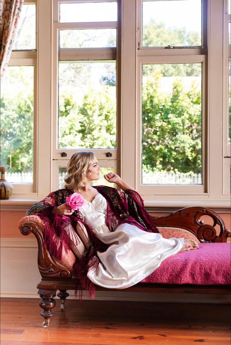 Op de afbeelding: Een vrouw in een witte satijnen jurk en een bordeauxrode shawl zit op een chaise longue met een roze roos in haar hand. Ze kijkt uit een raam met uitzicht op groene bomen.