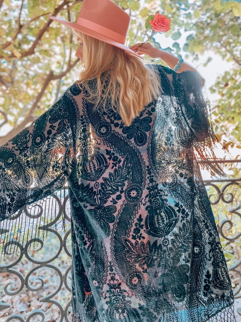 May include: A black sheer kimono with a floral pattern and fringe trim. The kimono is worn over a pink tank top. The person is wearing a pink hat and holding a pink rose.