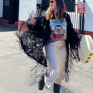 May include: A woman wearing a black lace kimono with fringe, a grey t-shirt with a truck graphic and the text "LOVE ROAD", a beige satin skirt, and black boots. She is holding a white suitcase with gold accents. The sign behind her reads "CAUTION WATCH OUT FOR CARS & PEDESTRIANS".