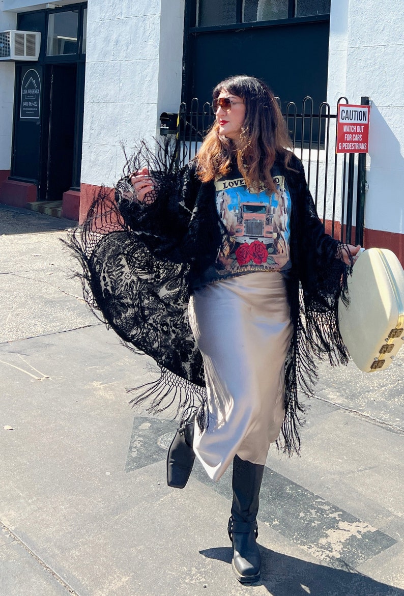 May include: A woman wearing a black lace kimono with fringe, a grey t-shirt with a truck graphic and the text "LOVE ROAD", a beige satin skirt, and black boots. She is holding a white suitcase with gold accents. The sign behind her reads "CAUTION WATCH OUT FOR CARS & PEDESTRIANS".