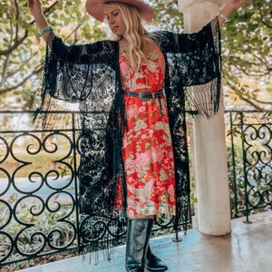 May include: A woman wearing a black lace kimono with fringe, a red floral midi dress, a pink cowboy hat, and black knee-high boots. She is holding a pink flower.