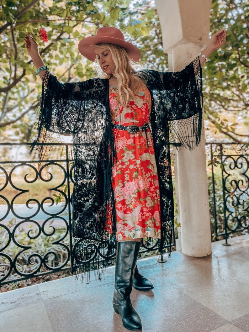 May include: A woman wearing a black lace kimono with fringe, a red floral midi dress, a pink cowboy hat, and black knee-high boots. She is holding a pink flower.