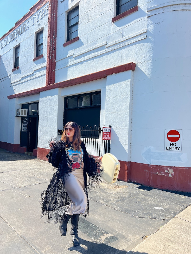 May include: A woman wearing a black fringed kimono over a graphic t-shirt and a beige skirt. She is carrying a white vintage suitcase and walking down a street with a red brick building in the background. The building has a sign that says "Beaurepaire Tyre".