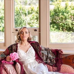 Op de afbeelding: Een vrouw in een witte satijnen jurk en een bordeauxrode en blauwe gebloemde shawl zit op een vintage bank. Ze houdt een roze roos in haar hand. De bank staat voor een raam met uitzicht op groene bomen.