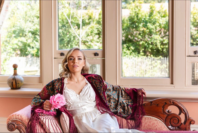 Op de afbeelding: Een vrouw in een witte satijnen jurk en een bordeauxrode en blauwe gebloemde shawl zit op een vintage bank. Ze houdt een roze roos in haar hand. De bank staat voor een raam met uitzicht op groene bomen.