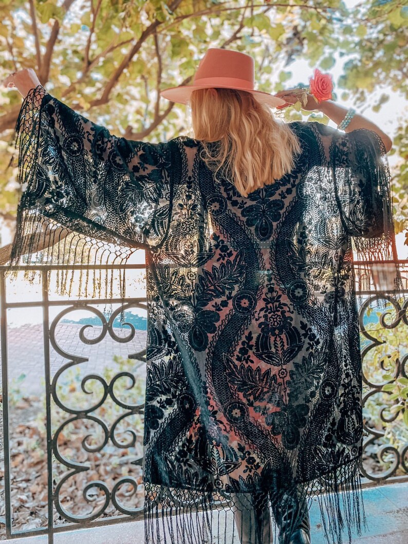 May include: A black sheer kimono with a floral pattern and fringe trim. The kimono is worn over a pink top and the person is wearing a pink hat.