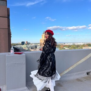 May include: A woman wearing a black lace dress with fringe, a red beret, and black shoes stands on a rooftop with a city skyline in the background.