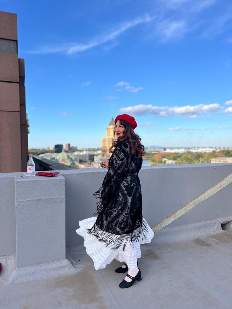 May include: A woman wearing a black lace dress with fringe, a red beret, and black shoes stands on a rooftop with a city skyline in the background.