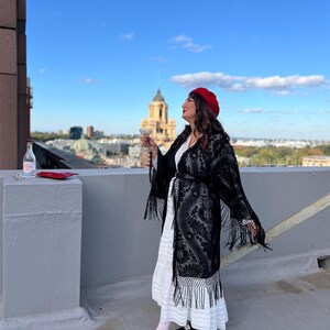 May include: A woman wearing a black lace kimono with fringe, a white dress, and a red beret, stands on a rooftop holding a glass of champagne and looking out at the city skyline.