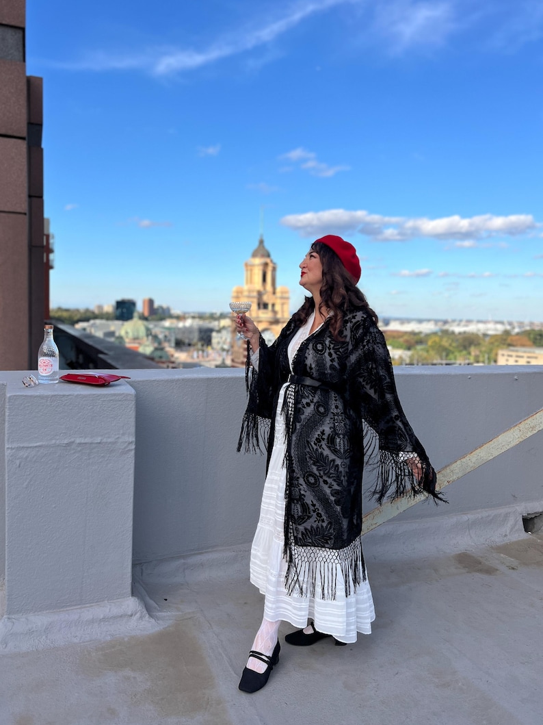 May include: A woman wearing a black lace kimono with fringe, a white dress, and a red beret, stands on a rooftop holding a glass of champagne and looking out at the city skyline.