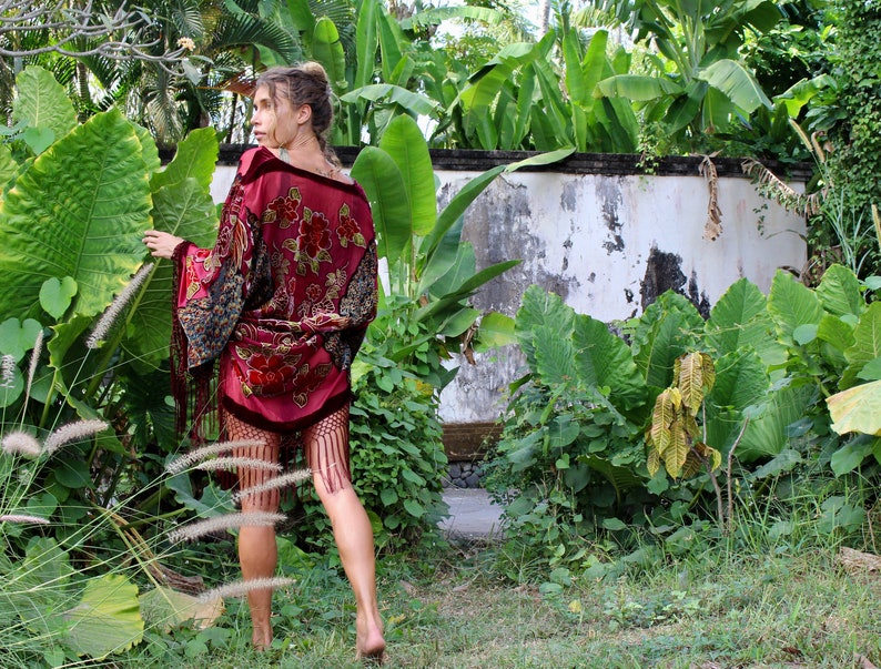 Op de afbeelding: Een vrouw draagt een rode fluwelen kimono met bloemenborduursel en franjes. De kimono heeft een zwart en gouden patroon op de mouwen. Ze staat in een tuin met weelderig groen gebladerte.