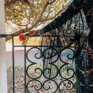 May include: A woman wearing a black lace kimono with a floral pattern, holding a single red rose in front of a wrought iron gate.