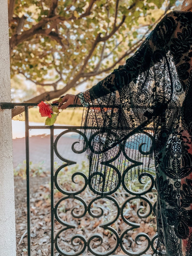 May include: A woman wearing a black lace kimono with a floral pattern, holding a single red rose in front of a wrought iron gate.