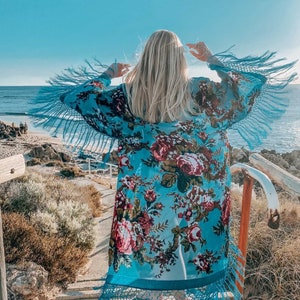 May include: A woman wearing a blue floral kimono with fringe trim. The kimono has a white background with large pink and red floral designs. The woman is standing on a wooden staircase with a view of the ocean in the background.
