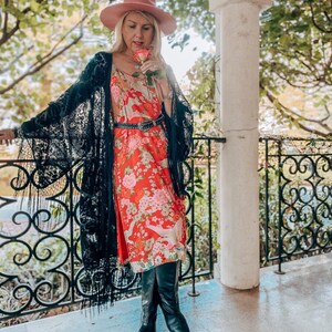 May include: A woman wearing a black lace kimono over a red floral dress with a black belt. She is holding a pink rose and wearing a pink hat and black boots.