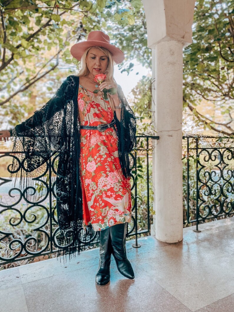 May include: A woman wearing a black lace kimono over a red floral dress with a black belt. She is holding a pink rose and wearing a pink hat and black boots.