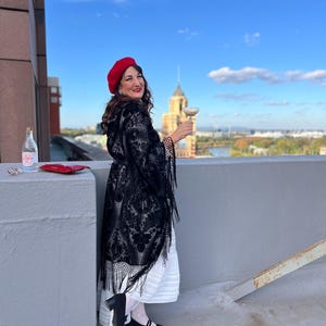 May include: A woman wearing a red beret, a black lace shawl, and a white dress, stands on a rooftop with a city skyline in the background. She is holding a glass of champagne and smiling.