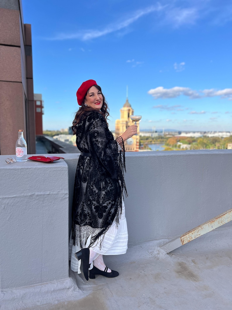 May include: A woman wearing a red beret, a black lace shawl, and a white dress, stands on a rooftop with a city skyline in the background. She is holding a glass of champagne and smiling.