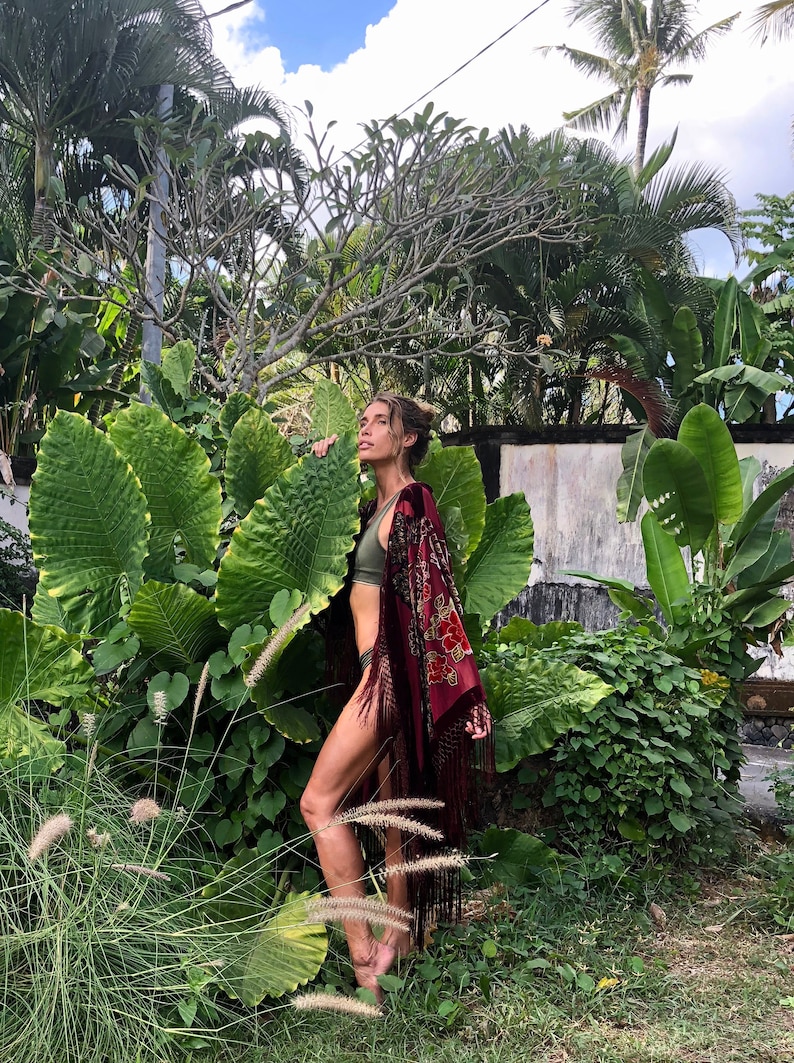 Op de afbeelding: Een vrouw in een rode fluwelen kimono met franjes en bloemenborduursel staat in een weelderige tropische tuin. Ze draagt een groene bikini top en heeft haar haar naar achteren getrokken.