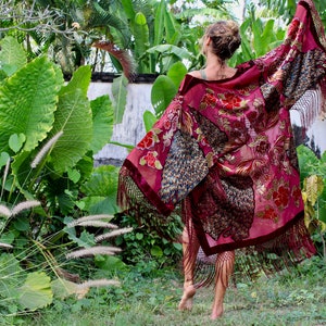 Op de afbeelding: Een vrouw draagt een lange, bordeauxrode kimono met gouden en pauwenveer borduurwerk en franjes. De kimono wappert in de wind terwijl ze door een weelderige tuin loopt.
