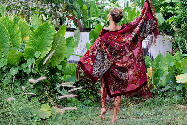 Op de afbeelding: Een vrouw draagt een lange, bordeauxrode kimono met gouden en pauwenveer borduurwerk en franjes. De kimono wappert in de wind terwijl ze door een weelderige tuin loopt.
