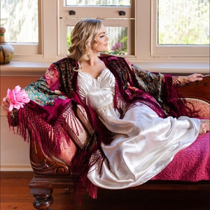Op de afbeelding: Een vrouw in een witte satijnen slipjurk en een bordeauxrode fluwelen sjaal met franjes zit op een vintage chaise longue. De sjaal heeft een pauwenmotief en is over haar schouders gedrapeerd. Ze houdt een roze bloem in haar hand.