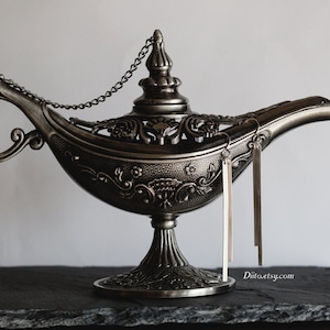 Pode incluir: Um par de brincos longos de franja prateada pendurados em uma lâmpada de gênio decorativa. A lâmpada é feita de metal e tem um design detalhado.