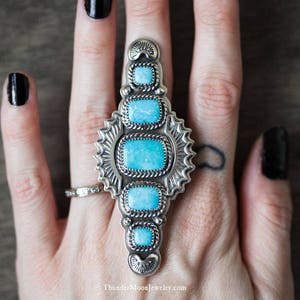 K&ouml;nnte beinhalten: Ein silberner Ring mit T&uuml;rkis-Steinen, die in einem geometrischen Design eingefasst sind. Der Ring wird an einer Hand mit schwarzem Nagellack getragen.