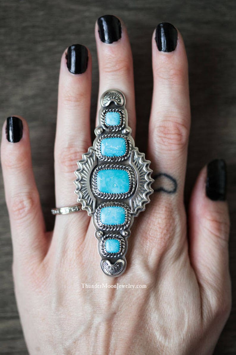 K&ouml;nnte beinhalten: Ein silberner Ring mit T&uuml;rkis-Steinen, die in einem geometrischen Design eingefasst sind. Der Ring wird an einer Hand mit schwarzem Nagellack getragen.