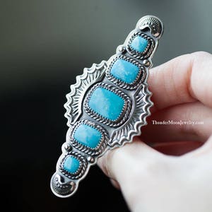 K&ouml;nnte beinhalten: Ein silberner Ring mit einem T&uuml;rkis-Stein-Inlay. Der Ring hat ein detailliertes Design mit einem Sonnenstrahlenmuster und einem seilartigen Rand um die Steine. Der Ring wird in einer Hand gehalten.