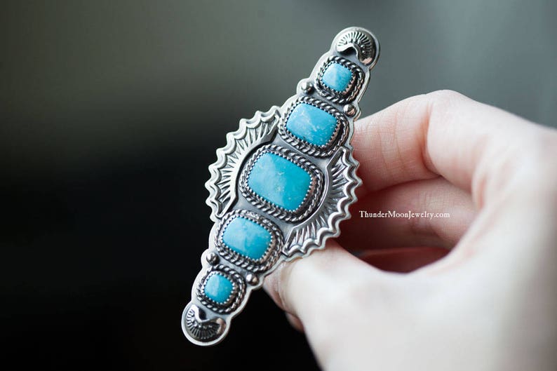 K&ouml;nnte beinhalten: Ein silberner Ring mit einem T&uuml;rkis-Stein-Inlay. Der Ring hat ein detailliertes Design mit einem Sonnenstrahlenmuster und einem seilartigen Rand um die Steine. Der Ring wird in einer Hand gehalten.