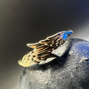 May include: A gold-coloured ring with a detailed wing design. The ring is on a dark grey surface.