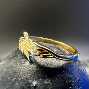 May include: A gold ring featuring a design of two hands clasped together, forming a heart shape. The ring is resting on a dark grey surface.