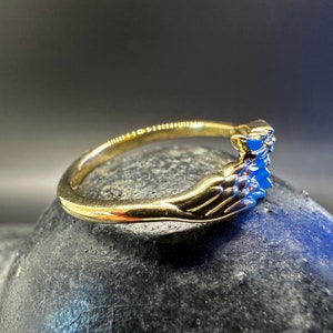 May include: A gold ring featuring two hands clasped together, representing a promise or commitment. The ring is set with blue gemstones.