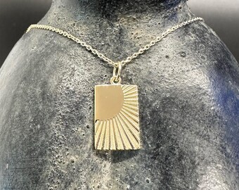 May include: A gold-colored pendant necklace featuring a rectangular pendant with a sunburst design. The pendant has a polished, half-circle sun and radiating lines. The necklace chain is delicate and silver-toned, creating a contrast with the pendant.