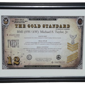 May include: A framed certificate with a gold seal and gold stripes. The certificate reads "The Gold Standard of Deckplate Leadership" and is awarded to BM1 (SW/AW) Michael S. Taylor, Jr. for 12 years of service. The certificate is dated August 8, 1988 and August 8, 2000.