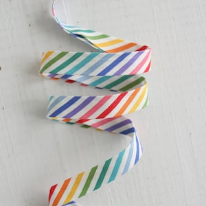 May include: A length of rainbow striped bias tape, with diagonal stripes in red, orange, yellow, green, blue, and purple. The tape is arranged in a flowing, curved pattern against a white background.