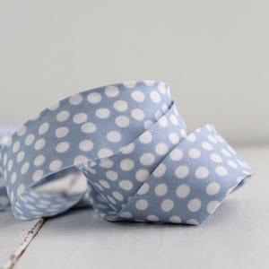 May include: A coiled length of light blue fabric ribbon featuring a repeating pattern of small, solid white polka dots. The ribbon is folded over itself, showing the consistent textile texture and the clear contrast between the blue and white colors.