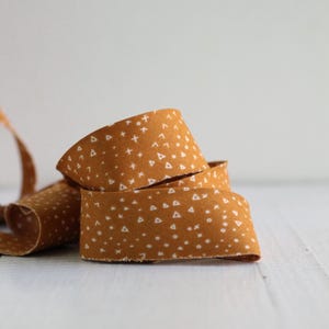 May include: A coiled length of mustard yellow fabric ribbon featuring a repeating pattern of small white geometric shapes, including tiny triangles, crosses, and circles, with raw, slightly frayed edges along the length of the textile strip.