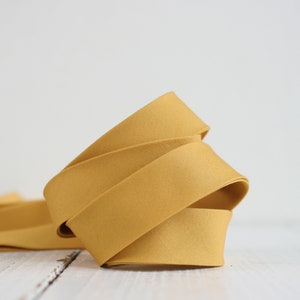 May include: A roll of mustard yellow fabric ribbon, folded and displayed on a white surface.
