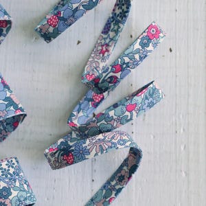 May include: A length of folded fabric bias tape featuring a dense floral pattern in shades of light blue, dusty pink, and white. The textile ribbon displays a repeating motif of small flowers, leaves, and berries across its entire surface.