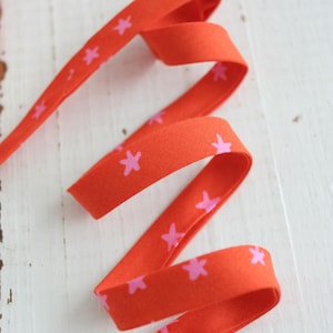 Puede incluir: Cinta naranja brillante con un patrón repetido de pequeñas estrellas rosas. La cinta está doblada y curvada, creando una forma dinámica y fluida. Esta cinta decorativa es adecuada para manualidades, envolver regalos u otros proyectos creativos.