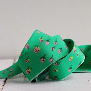 May include: A coiled length of green fabric ribbon featuring a repeating pattern of small pink strawberries with green stems and black seeds. The ribbon has a smooth texture and is displayed in a loose, overlapping loop to show the print.